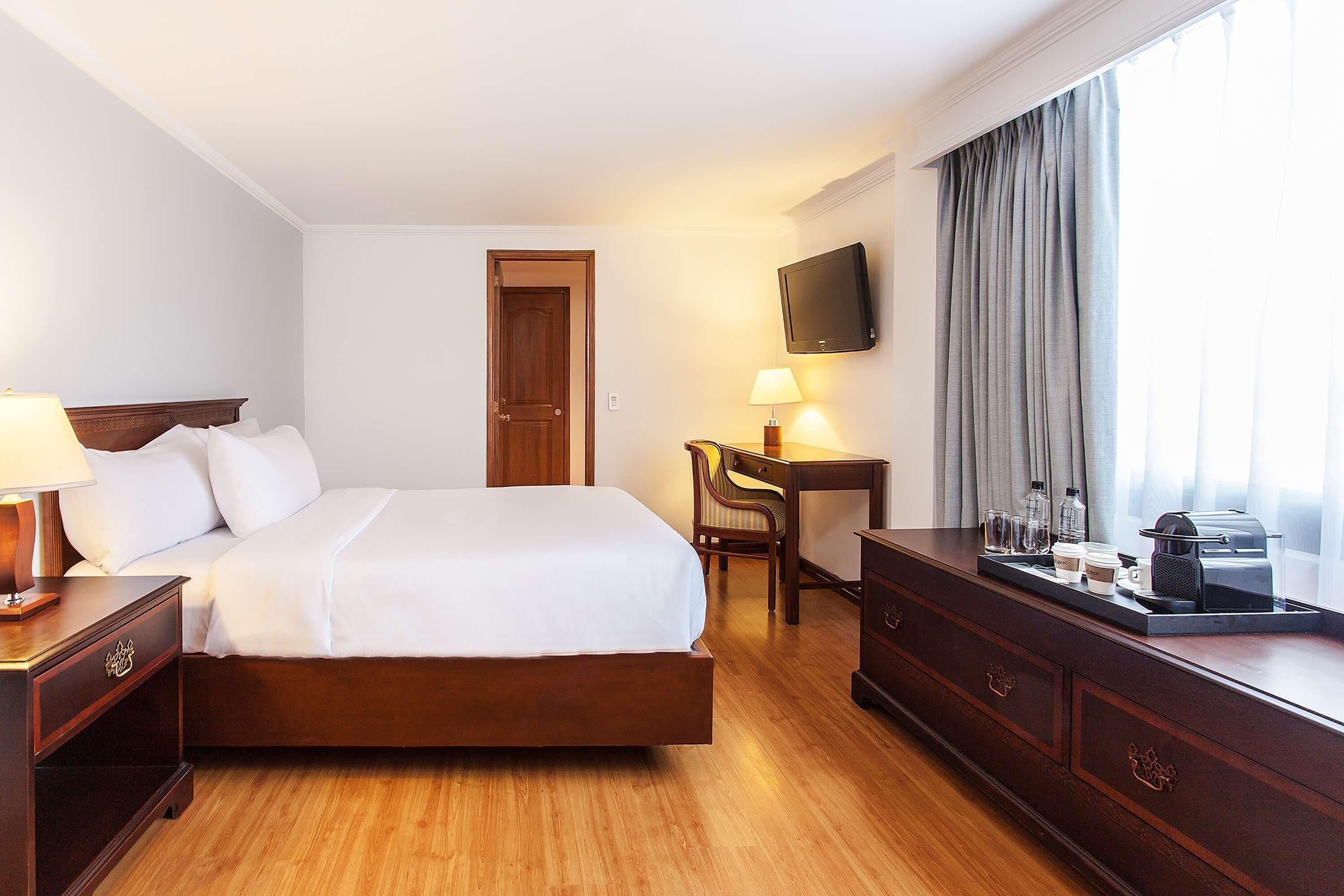 A hotel room with a bed, bedside table, lamp, desk, chair, TV, and window with curtains.