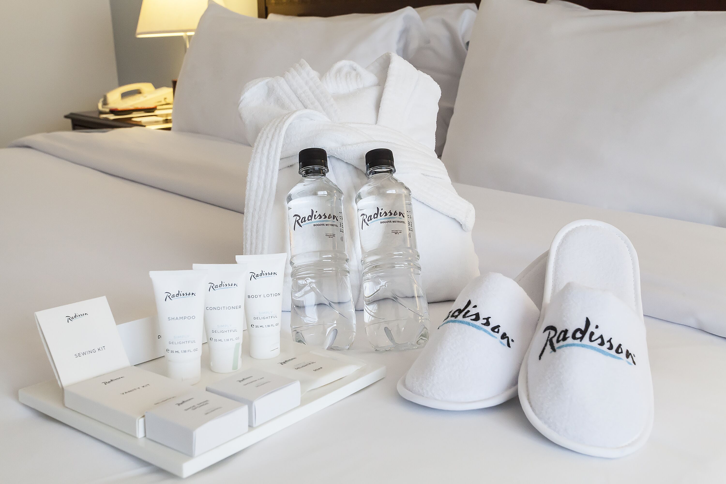 Ein ordentlich eingerichtetes Hotelzimmer mit einem Bett, einem Nachttisch mit Lampe sowie einem Set an Radisson-Toilettenartikeln, darunter Shampoo, Conditioner und Körperlotion.