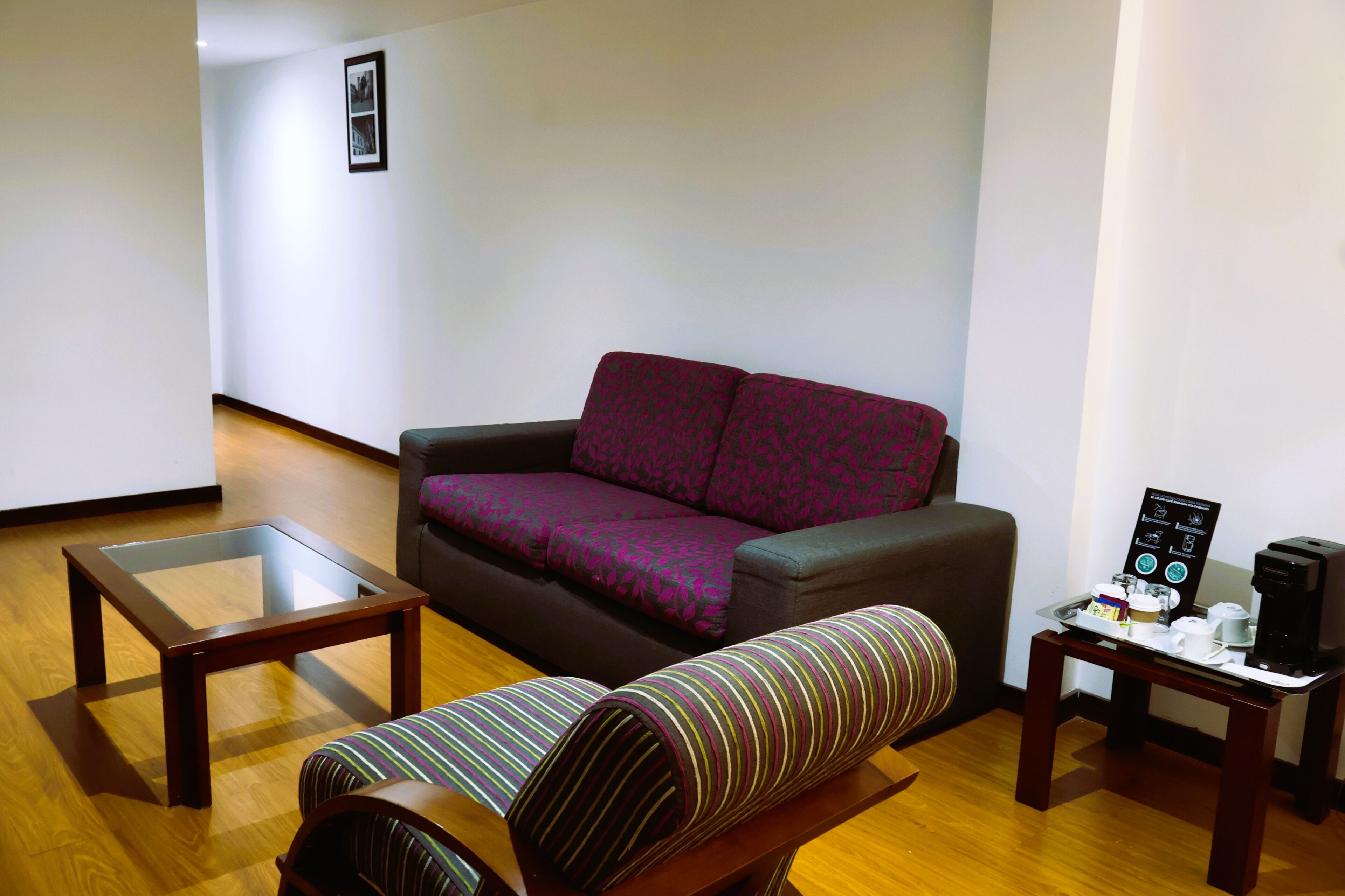 Una sala de estar con un sofá, un reposapiés a rayas, una mesa de centro con superficie de vidrio y una mesita con cafetera.