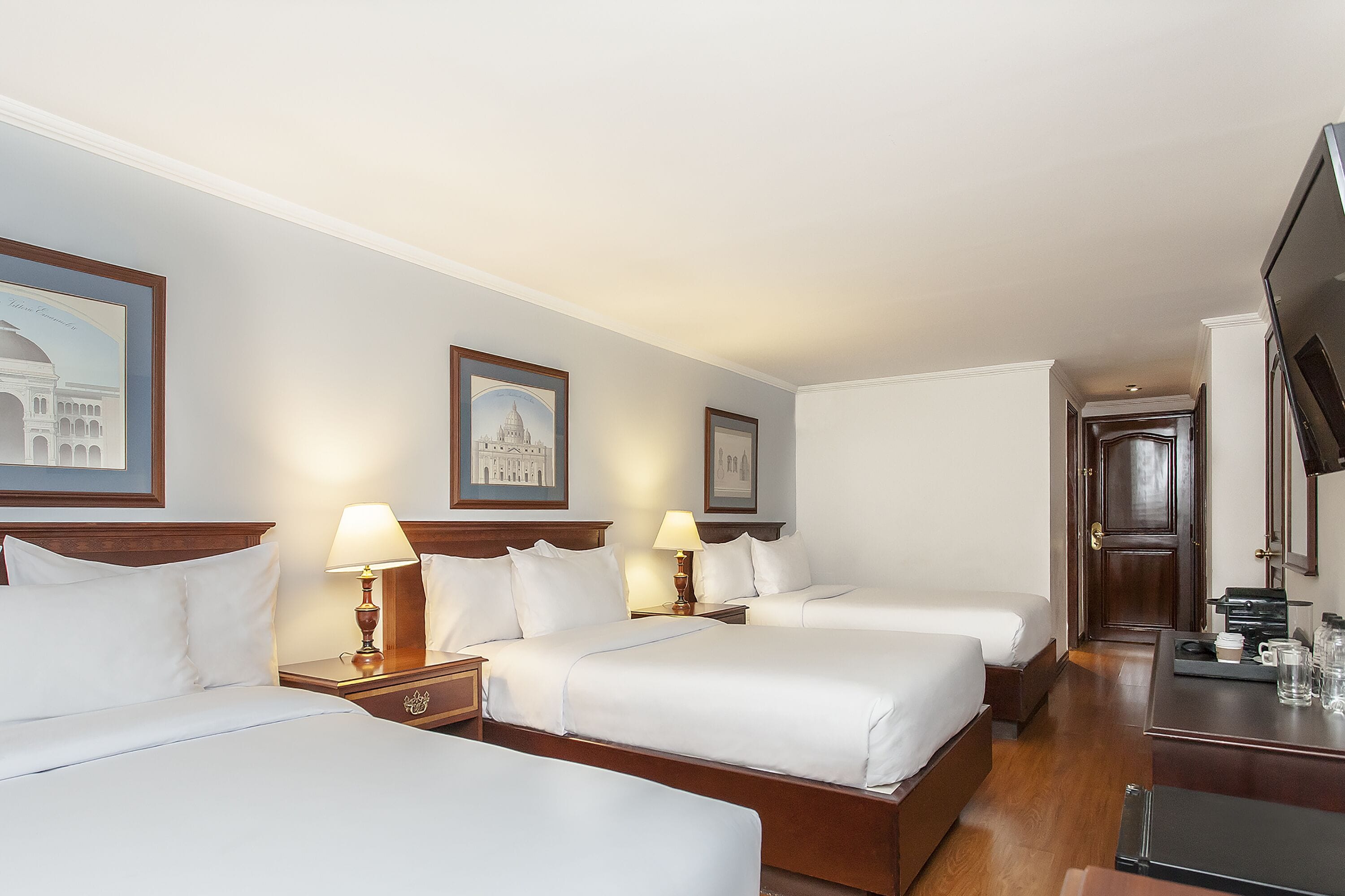 A hotel room with three beds, wooden headboards, white linens, and framed pictures on the walls.