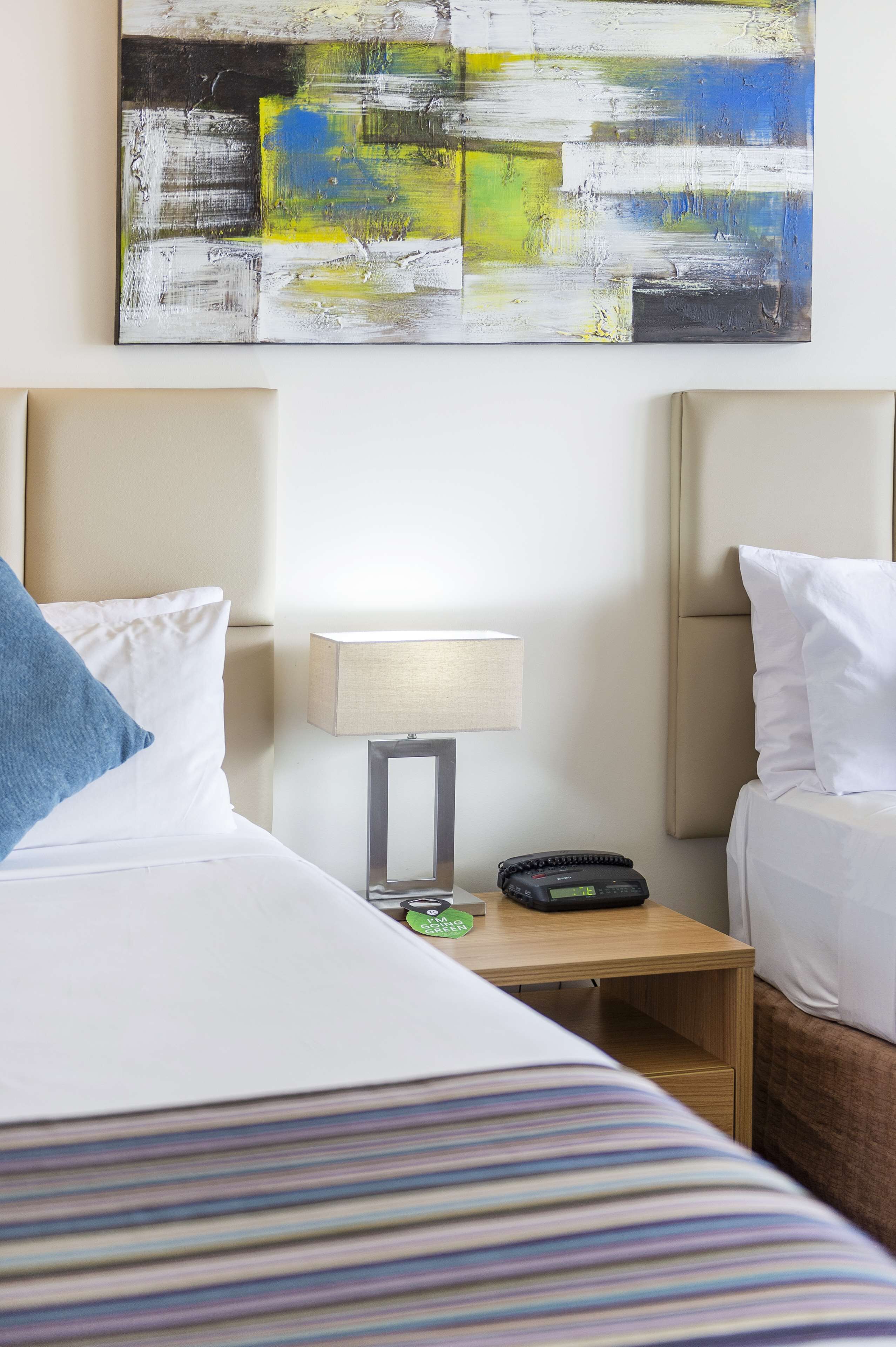 A hotel room with a bed, a bedside table with a lamp, a phone, and a wall art piece.