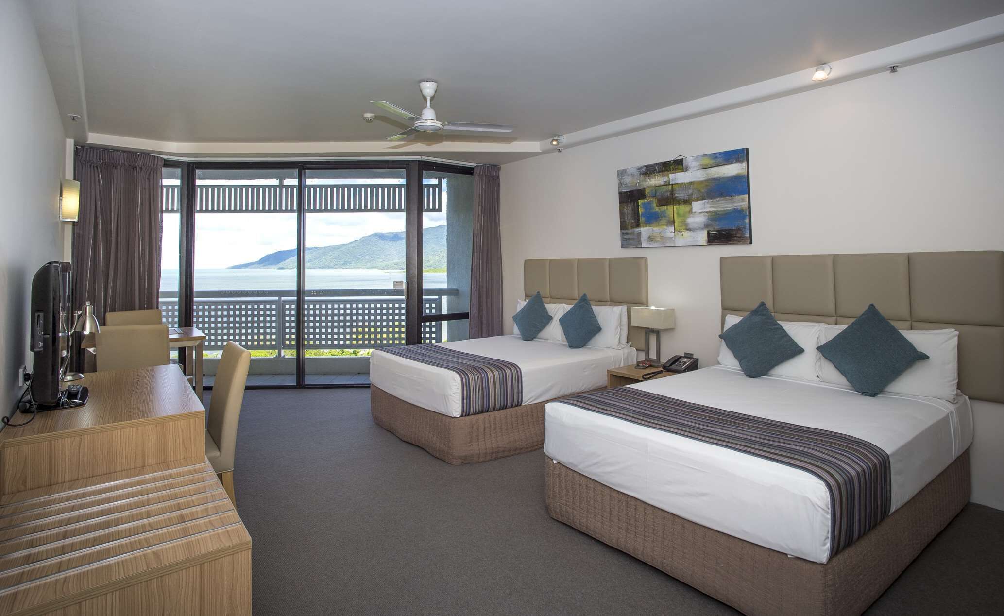 A hotel room with two beds, a desk, a chair, a ceiling fan, and a view of the sea.