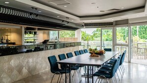 Un comedor amplio con una mesa larga y sillas azules, una zona de cocina con mostrador y estanterías, y ventanas grandes que ofrecen vistas a zonas verdes.