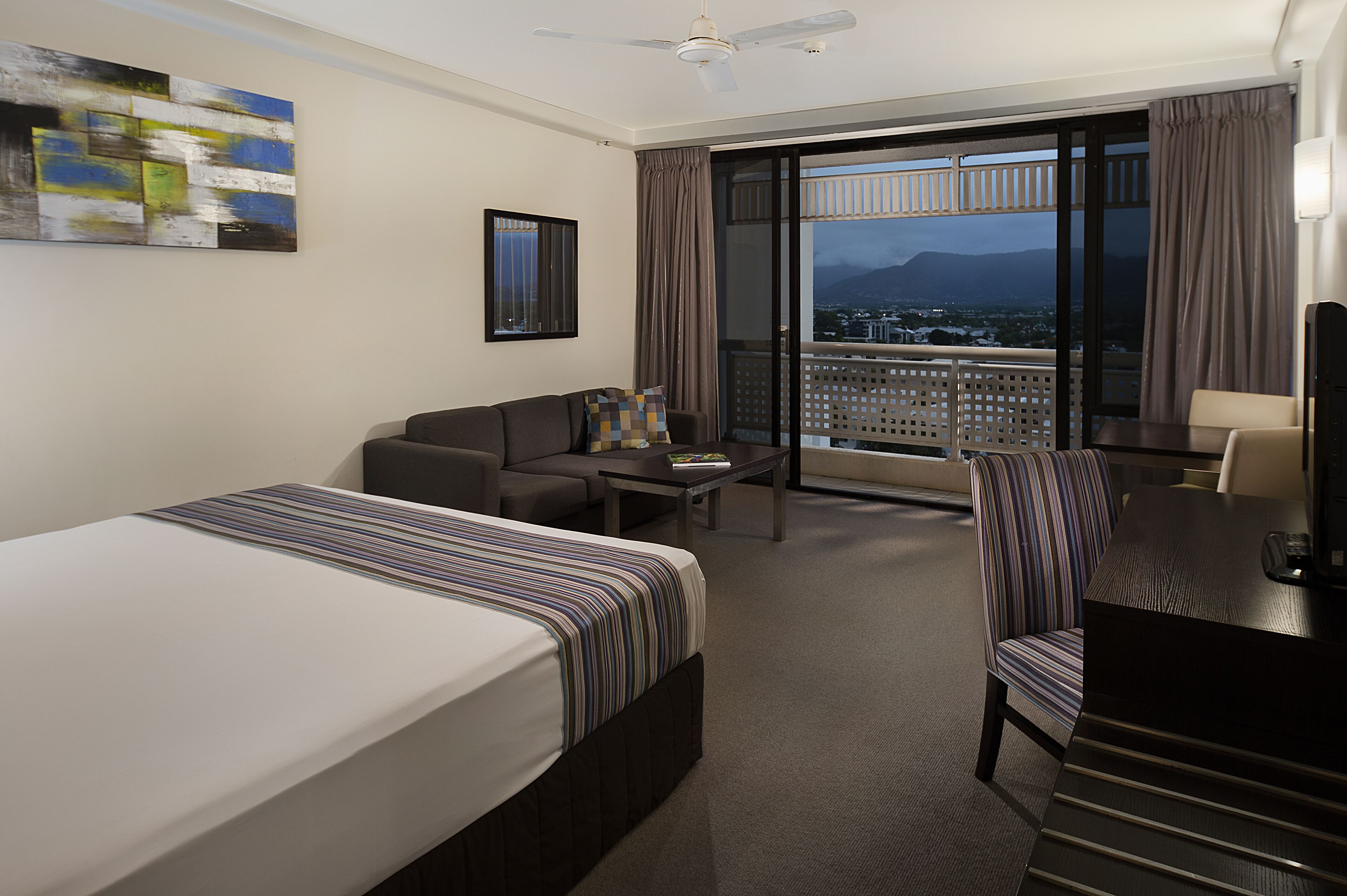 A hotel room with a bed, a sofa, a desk, a chair, a coffee table, and a large window with a view of the city and mountains.
