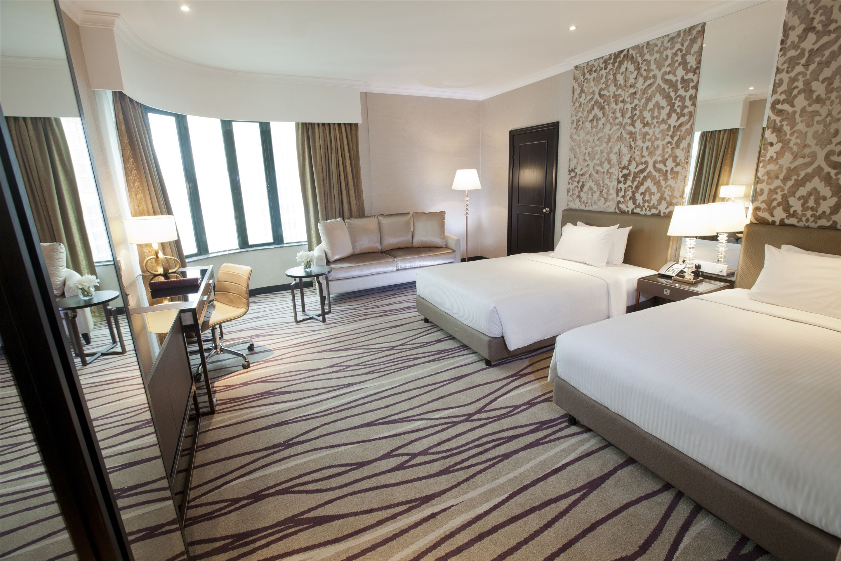 Habitación de hotel con dos camas, un escritorio con silla, un sofá y un ventanal con cortinas.