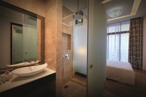 A modern bathroom with a large sink, a shower, and a bed in the adjacent room.