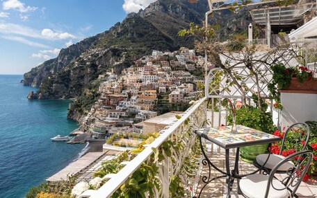 A balcony with a table and chair overlooking a coastal town with colorful buildings and a clear blue sea.