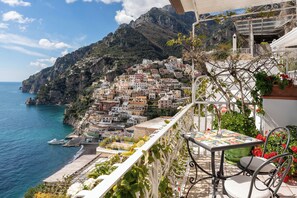 A balcony with a table and chair overlooking a coastal town with colorful buildings and a clear blue sea. - Hotel Eden Roc (Positano)