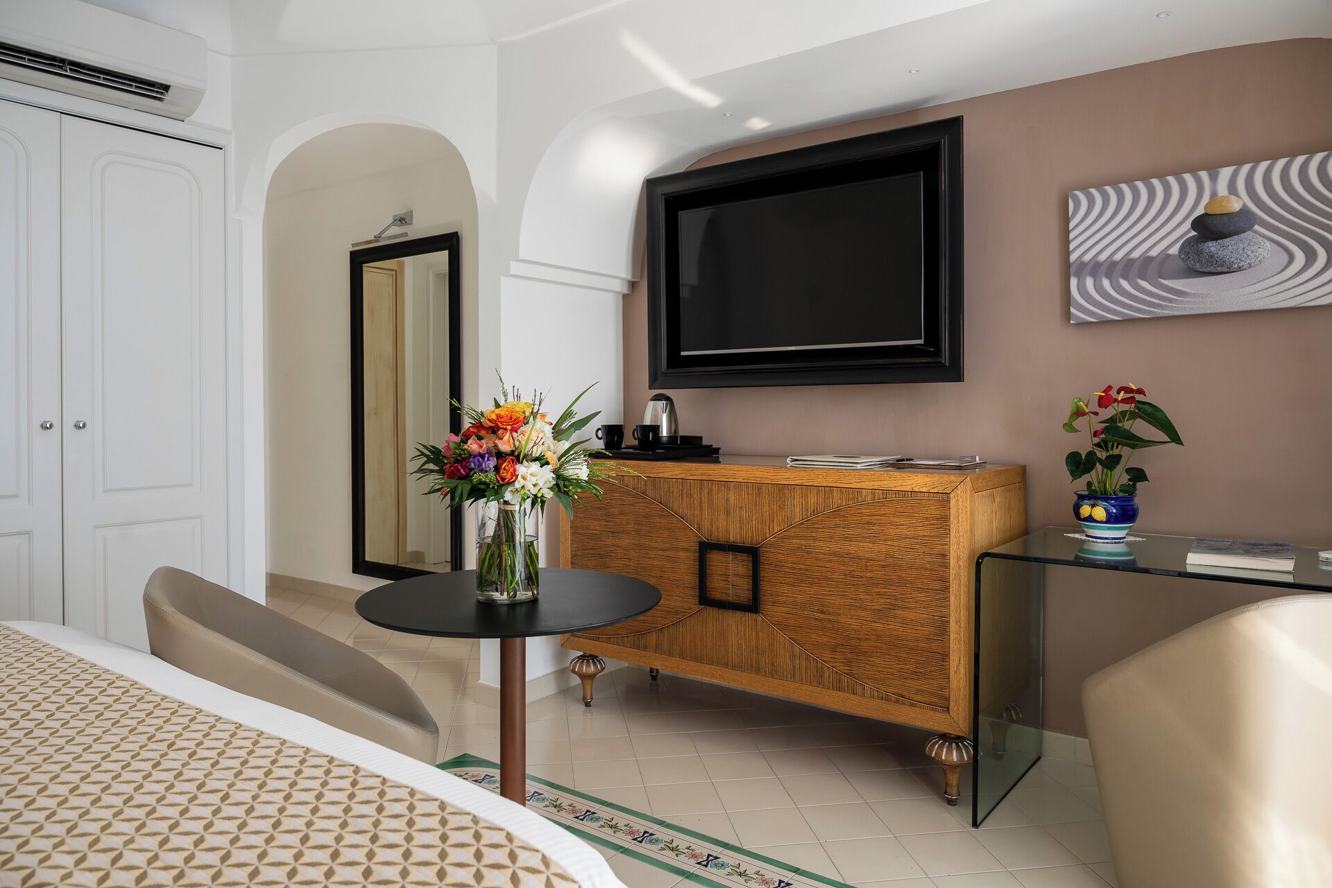 Une chambre d’hôtel moderne dotée d’un grand téléviseur à écran plat fixé au mur, d’une armoire en bois avec un plateau en verre, d’une petite table ronde agrémentée d’un vase de fleurs et d’un lit avec une literie à motifs.