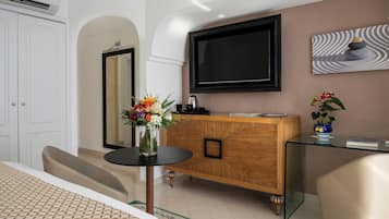 A modern hotel room with a large flat-screen TV mounted on the wall, a wooden cabinet with a glass top, a small round table with a vase of flowers, and a bed with patterned bedding.
