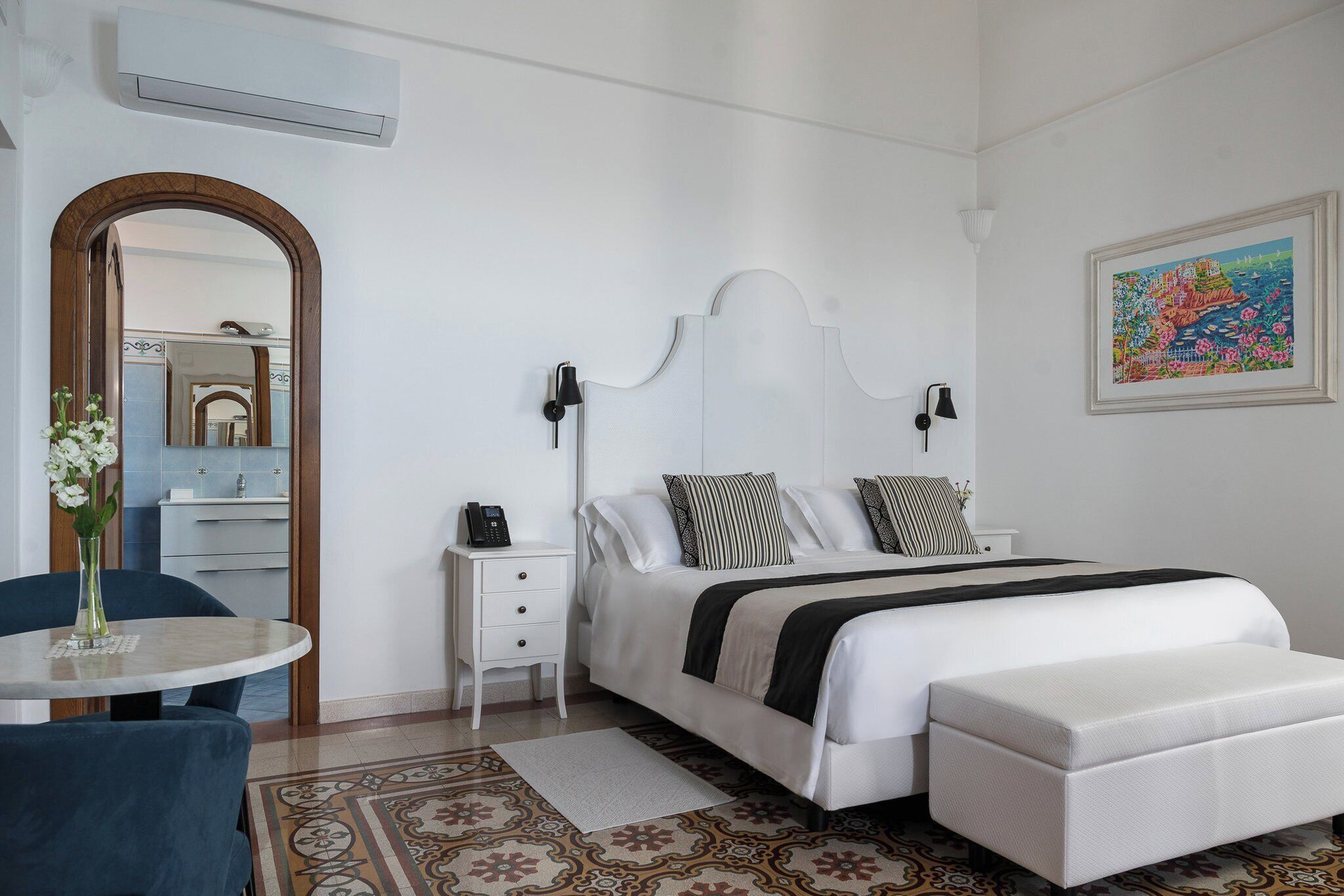A modern bedroom with a white bed, bedside lamps, a bedside table with a phone, a small round table with a vase, and a chair.