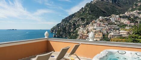 A balcony with lounge chairs and a hot tub overlooking a coastal town with buildings on a hillside.
