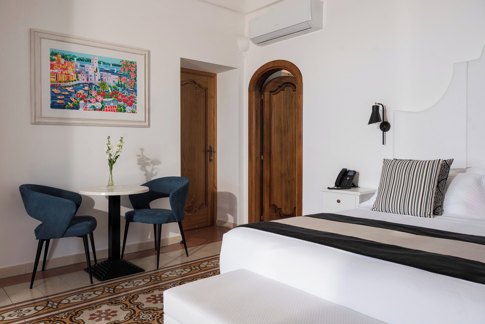 Habitación de hotel con una cama, un escritorio con dos sillas, una mesita con un florero, un cuadro en la pared y una alfombra estampada.