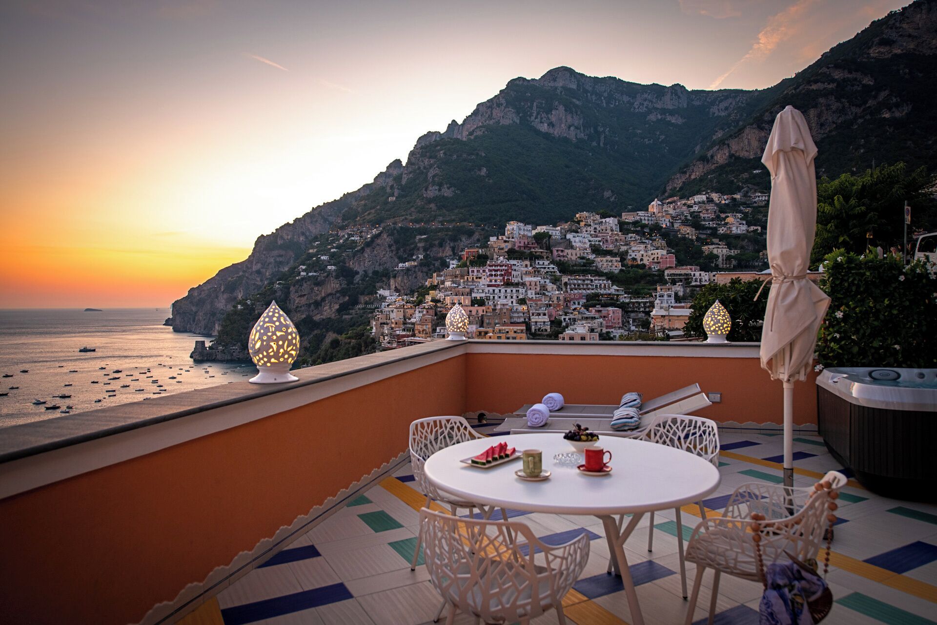 A terrace with a round table, chairs, and a view of a coastal town at sunset.
