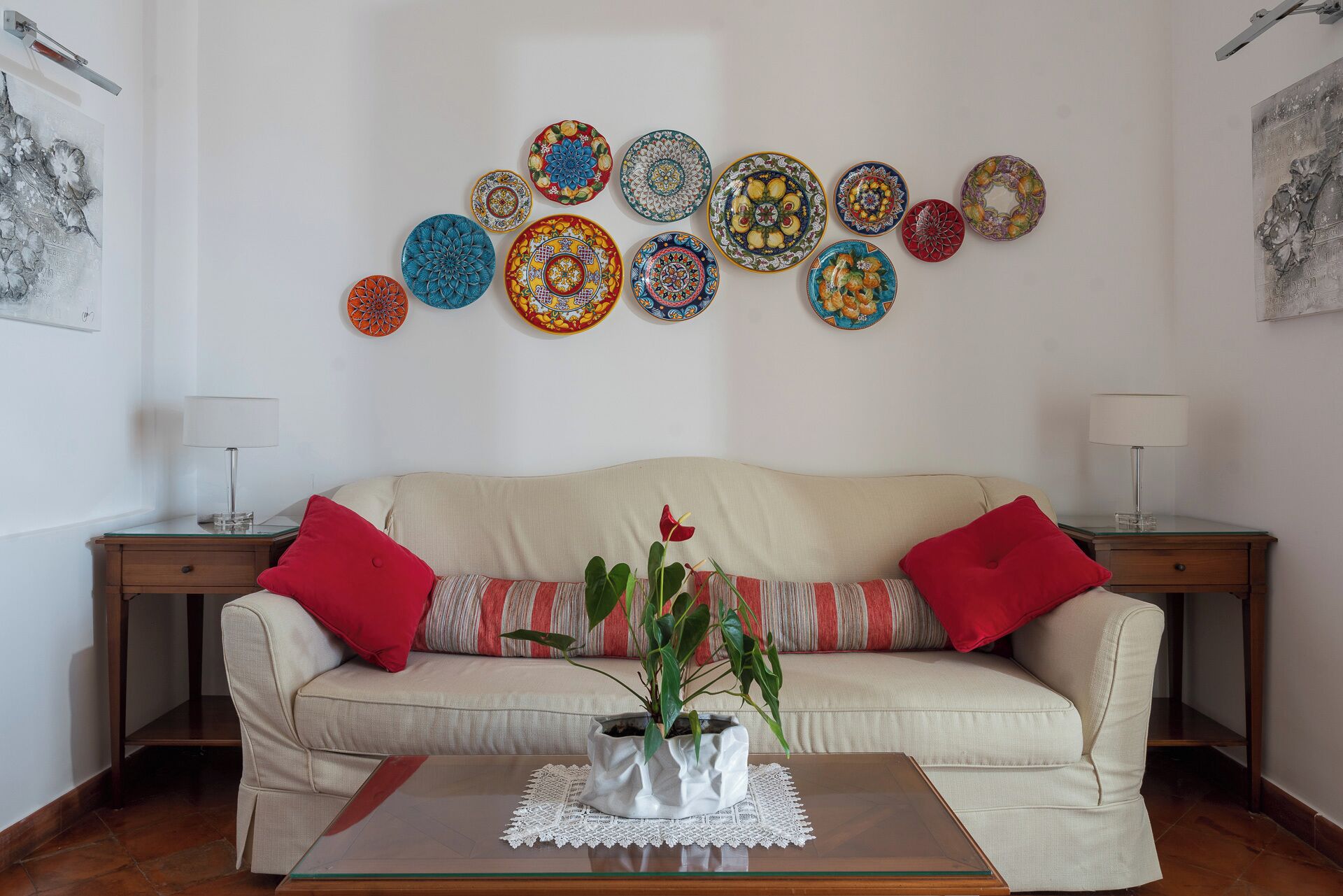 A living room with a beige sofa, red cushions, a glass coffee table with a white vase and plant, and a wall decorated with colorful plates.