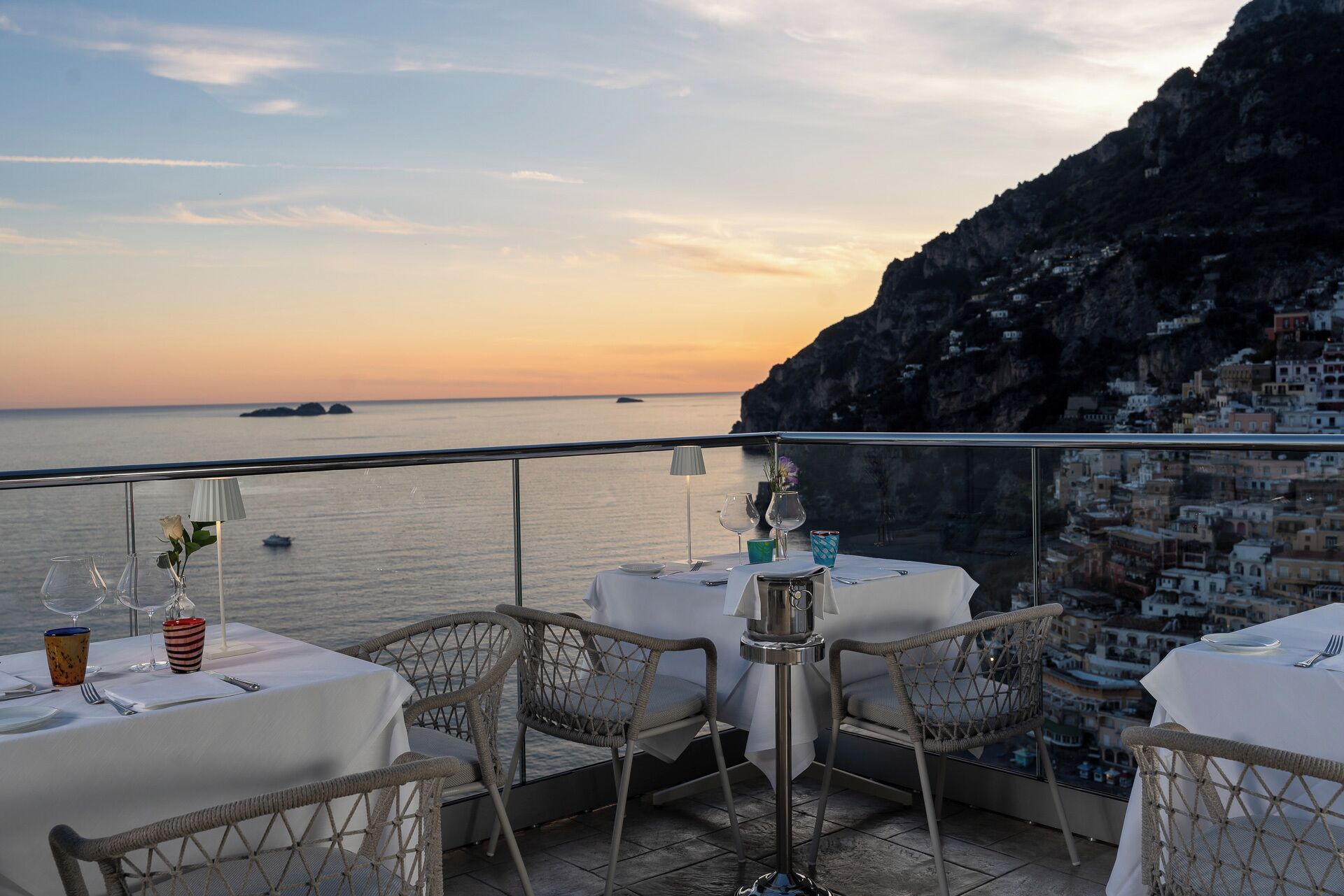 A terrace with tables set for dining, overlooking a coastal view with buildings and cliffs.