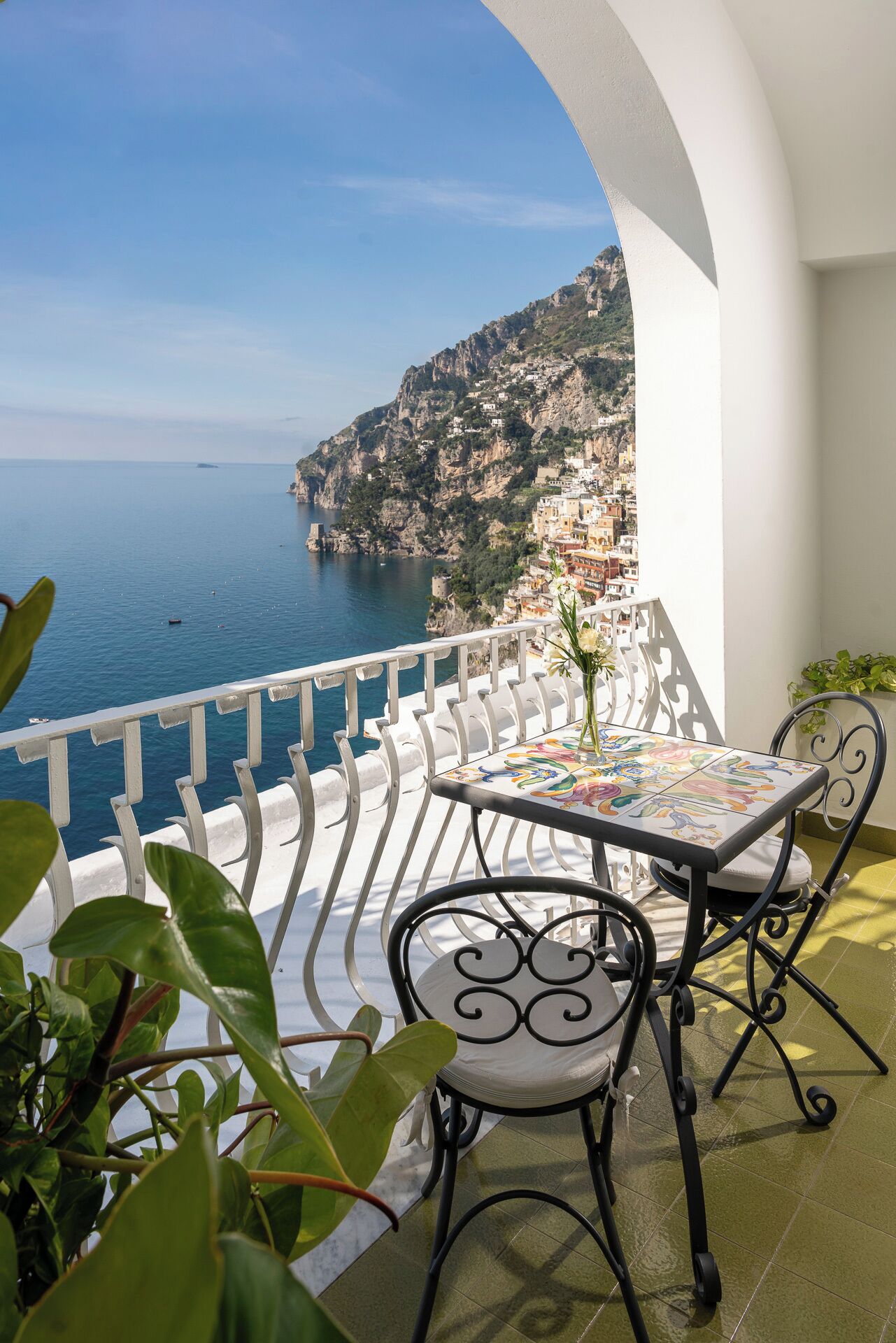 A balcony with a table and chairs, overlooking a coastal town and the sea.