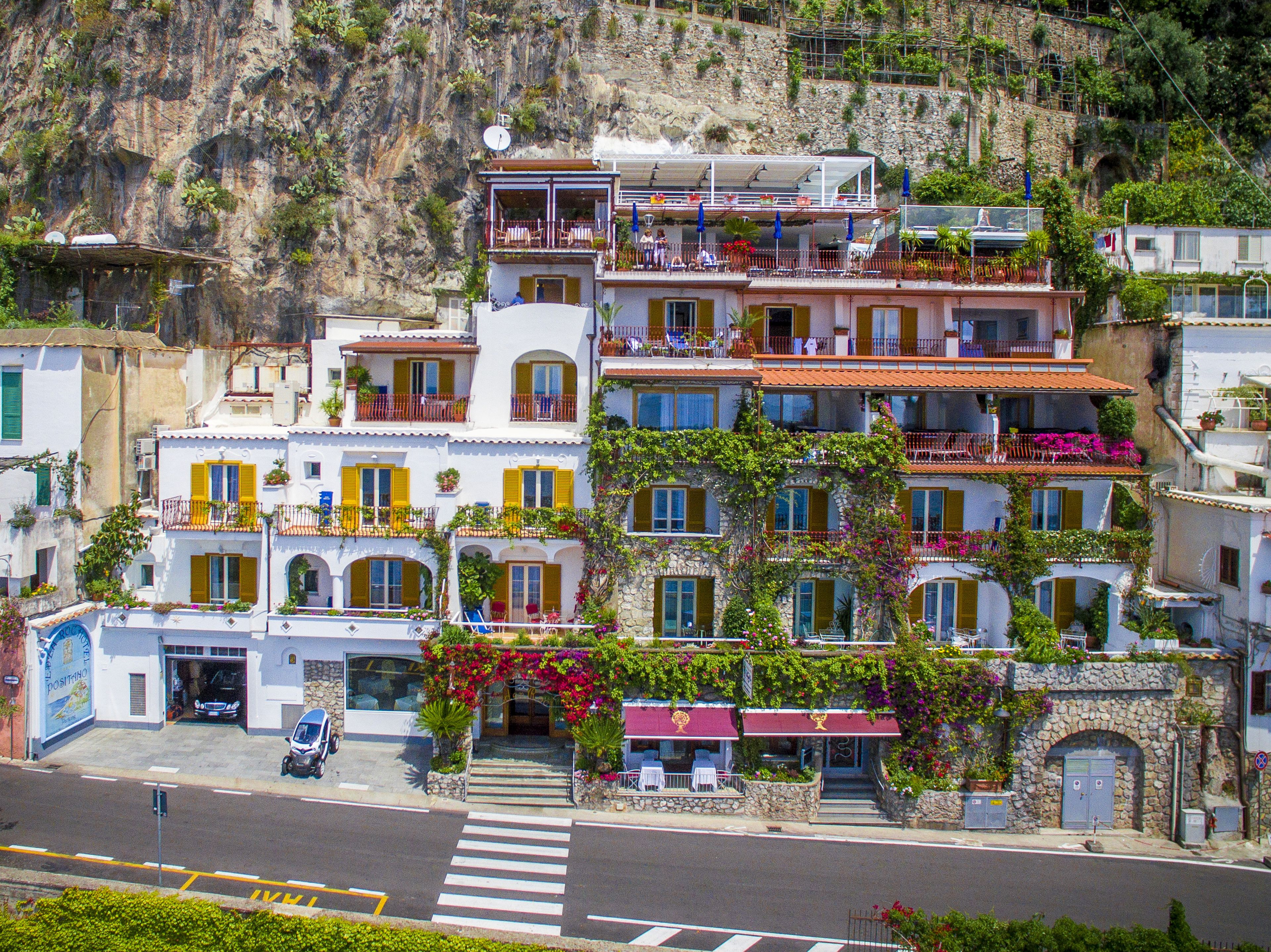 Un edifici de diversos pisos amb balcons, un carrer al davant i un paisatge de muntanyes al fons.