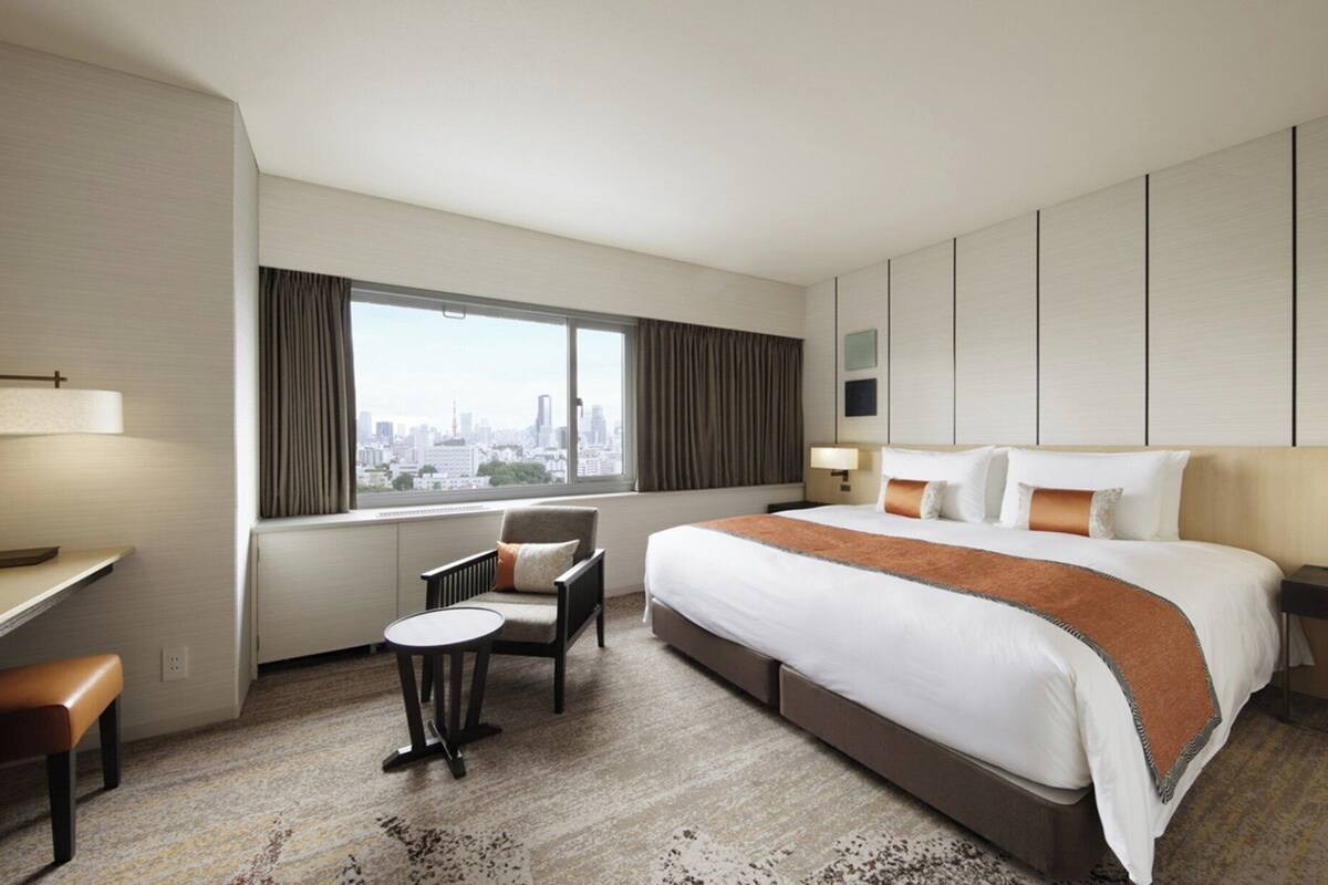 A hotel room with a large bed, a desk, a chair, and a view of a cityscape through a window.