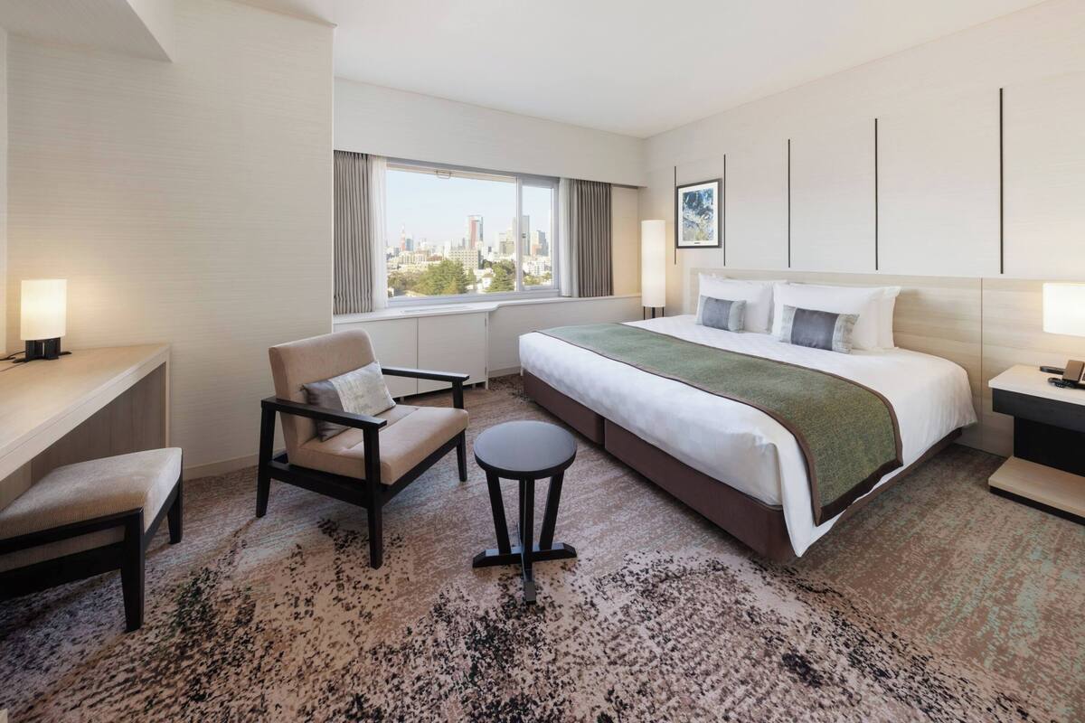 A hotel room with a bed, a desk, a chair, a small table, and a view of the city.