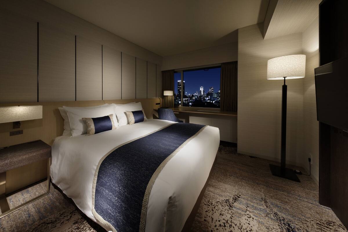 A hotel room with a large bed, a nightstand, a lamp, and a view of the city skyline.