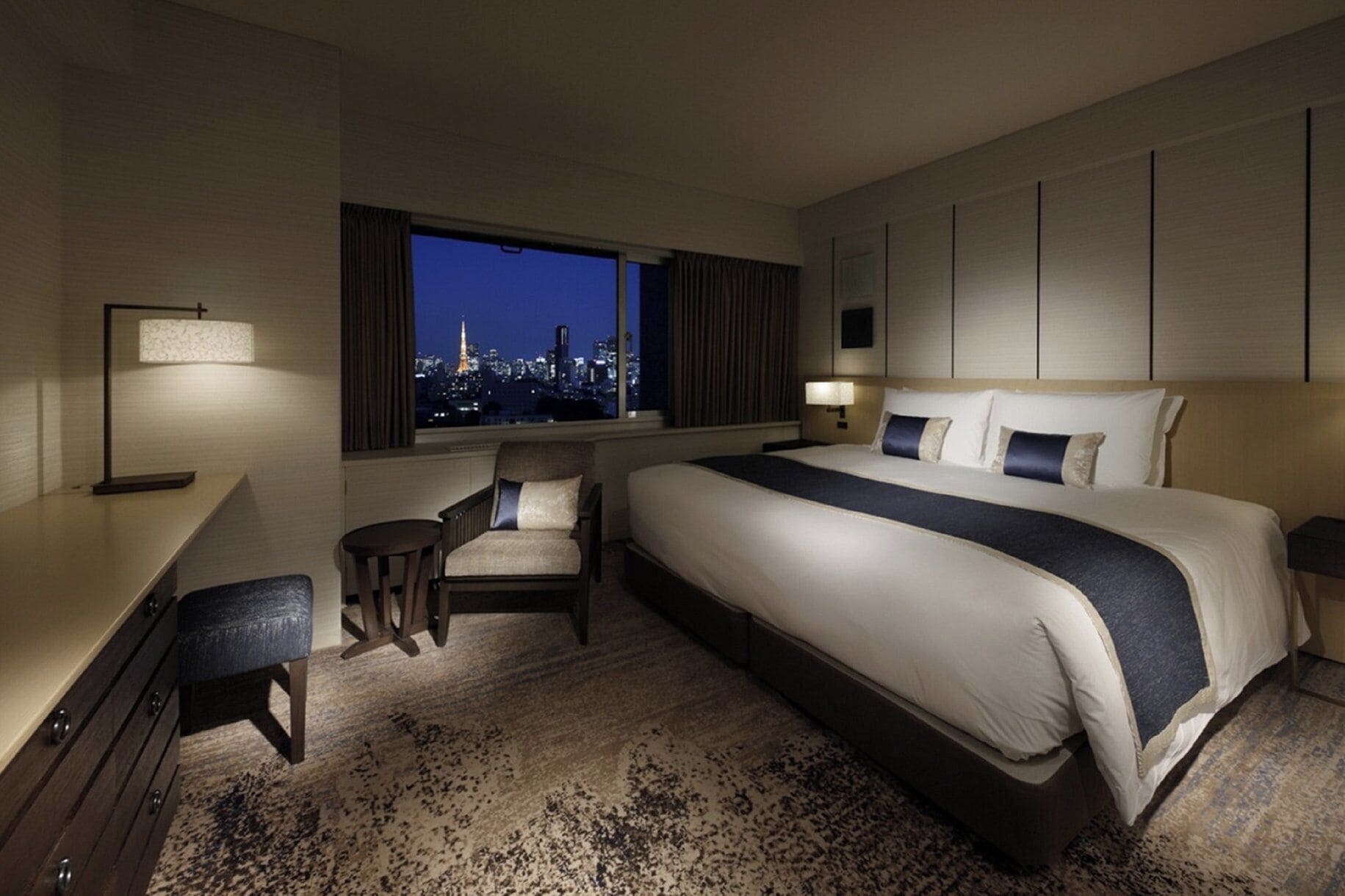 A hotel room with a large bed, a desk, a chair, and a view of a cityscape at night.
