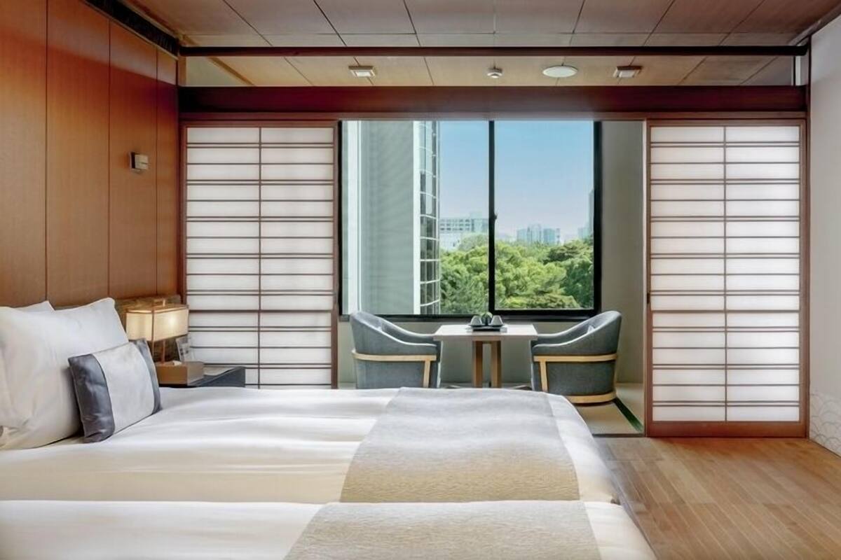 A hotel room with a large bed, a small table, two chairs, and a view of greenery and buildings.