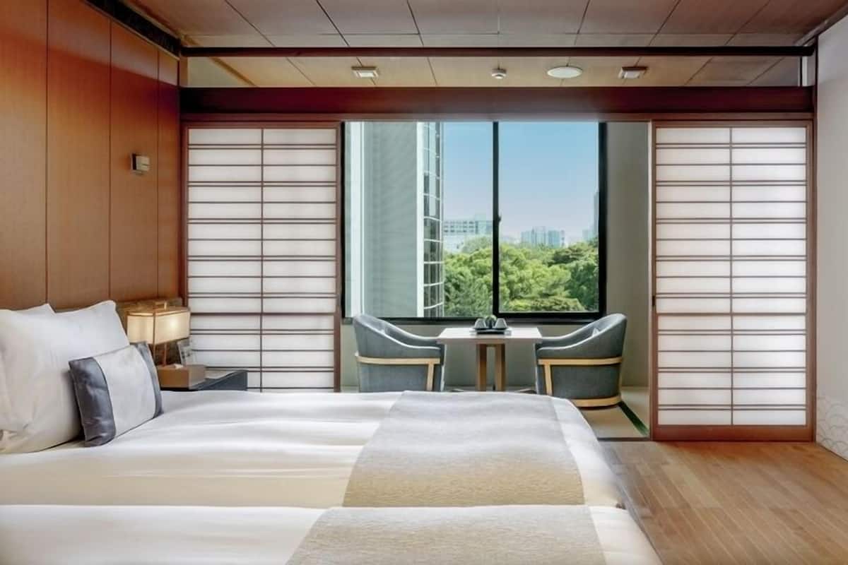 Habitación de hotel con una cama grande, una mesita, dos sillas y vistas a zonas verdes y edificios.