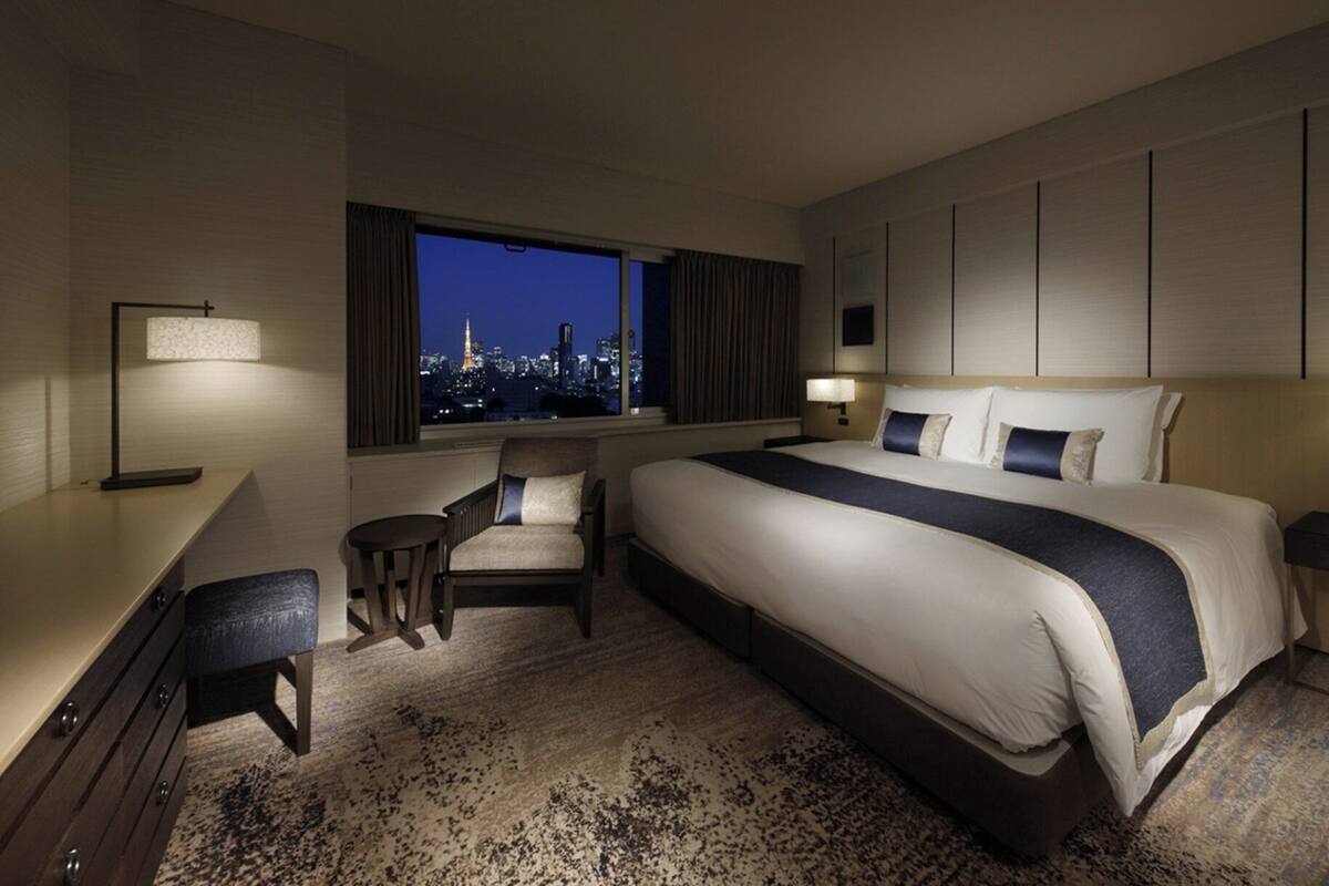 A hotel room with a large bed, a desk, a chair, and a view of a cityscape at night.