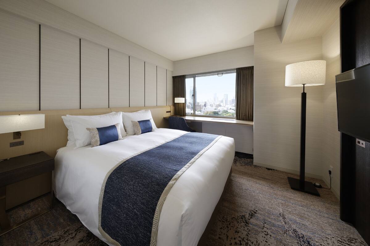 A hotel room with a large bed, a desk, a chair, a TV, and a window with a city view.