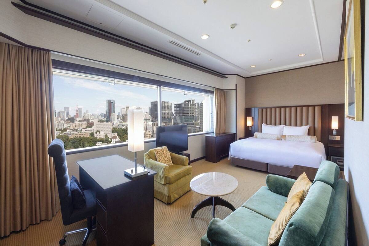 A hotel room with a large bed, a desk, a green sofa, a small round table, and a view of a cityscape through large windows.
