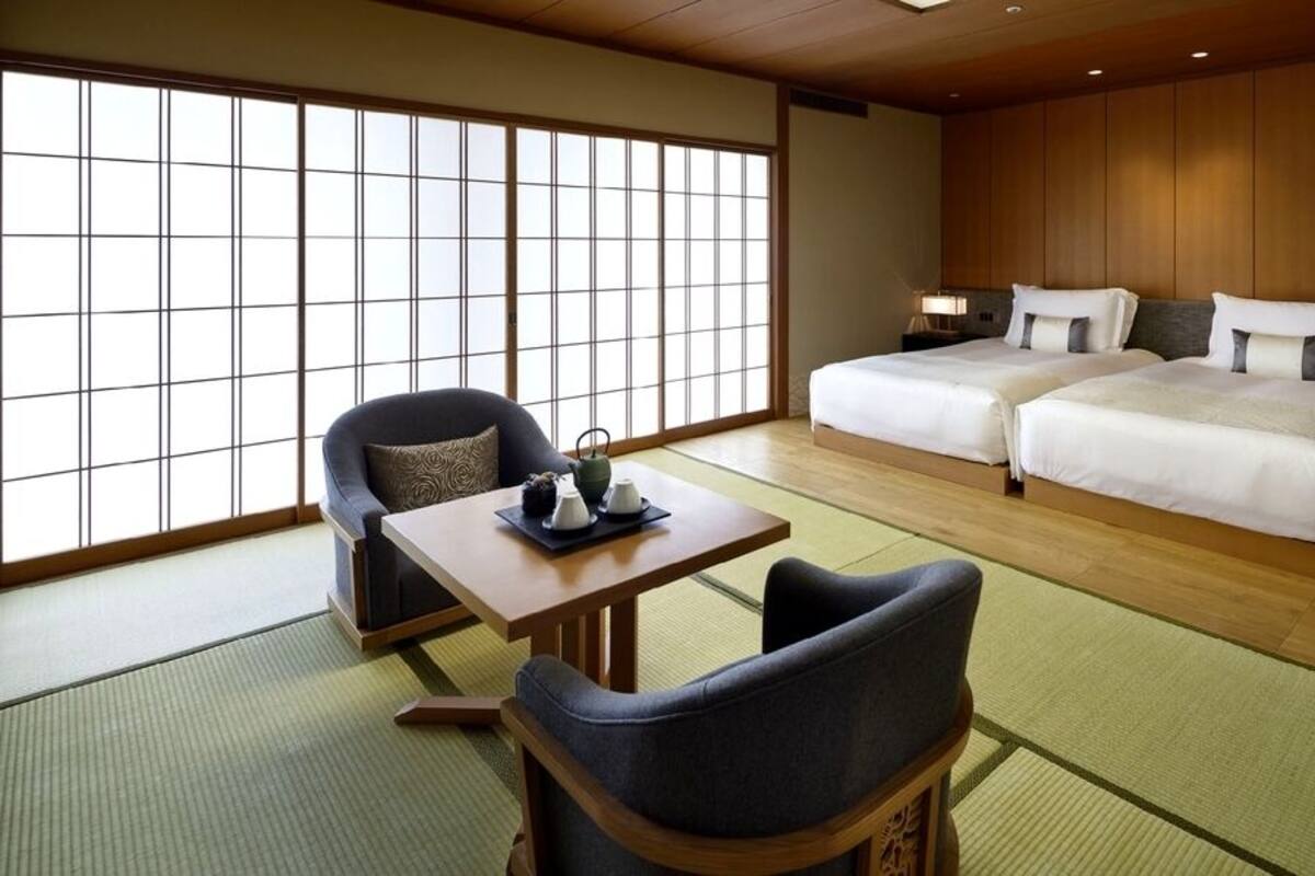 A traditional Japanese-style hotel room with tatami flooring, a low wooden bed, a tatami mat table, and a sliding shoji screen.