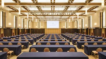 Ein Konferenzraum mit Reihen von Tischen und Stühlen, einer großen Leinwand im Vordergrund und einem modernen Deckenstil.
