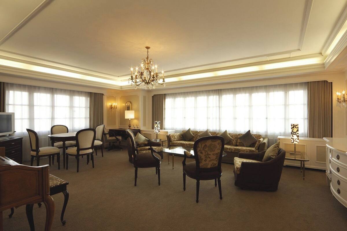A spacious hotel living room with a sofa set, chairs, a dining table, and a chandelier.