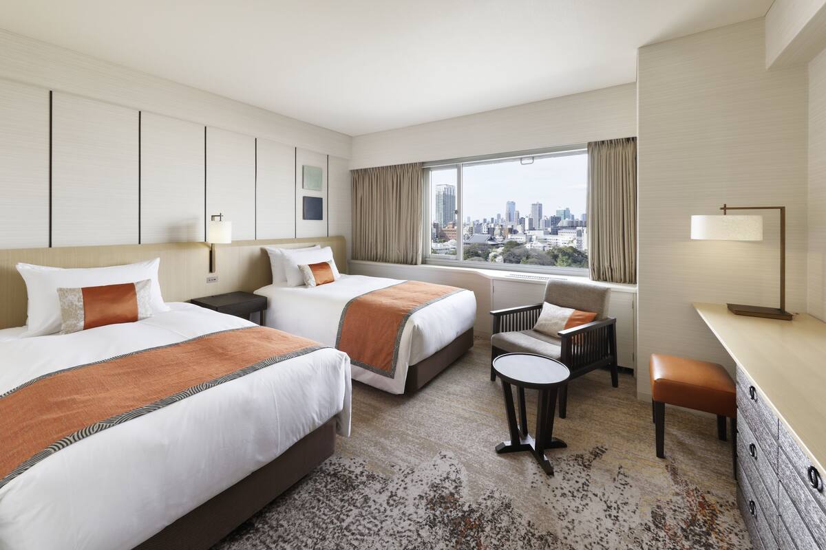 A hotel room with two beds, a desk, a chair, and a view of the city skyline.