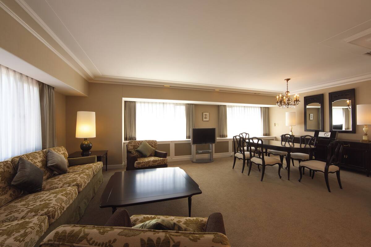 A hotel room with a sofa, armchair, dining table, chairs, a TV, and a chandelier.