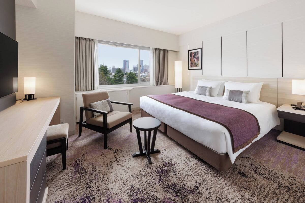 A hotel room with a bed, a chair, a desk, a TV, a window with a city view, and a lamp.
