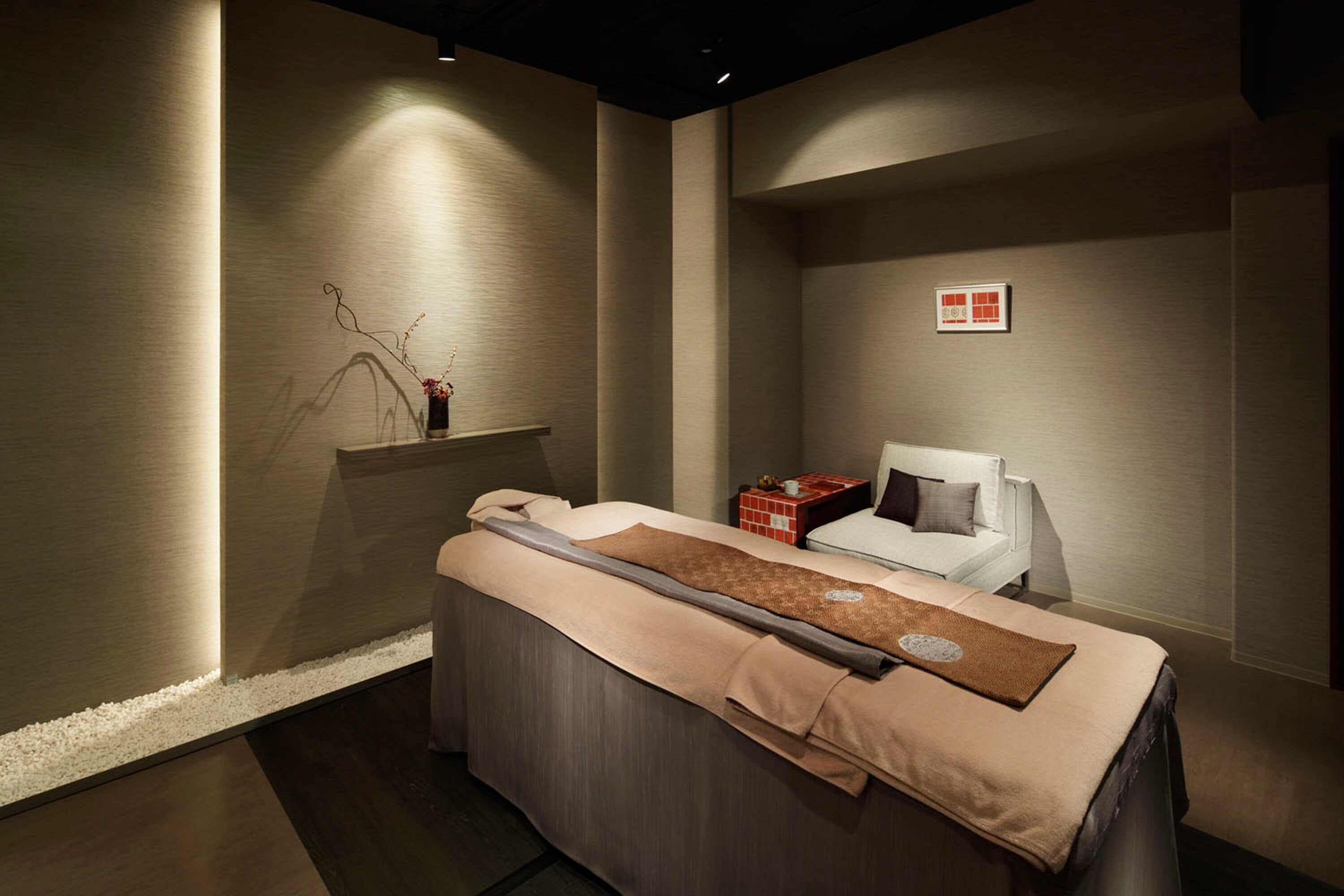 Un moderno cuarto de spa con una camilla de masaje, un estante pequeño con objetos decorativos y una silla blanca.