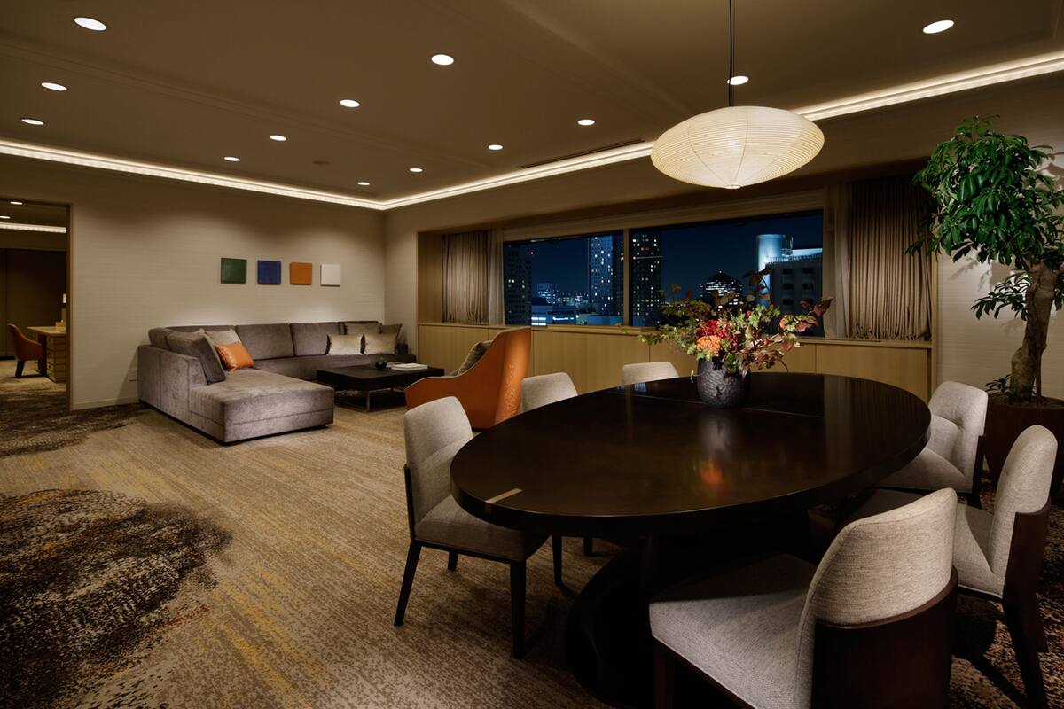 A modern hotel room with a dining area, a sofa, and a large window with a city view.