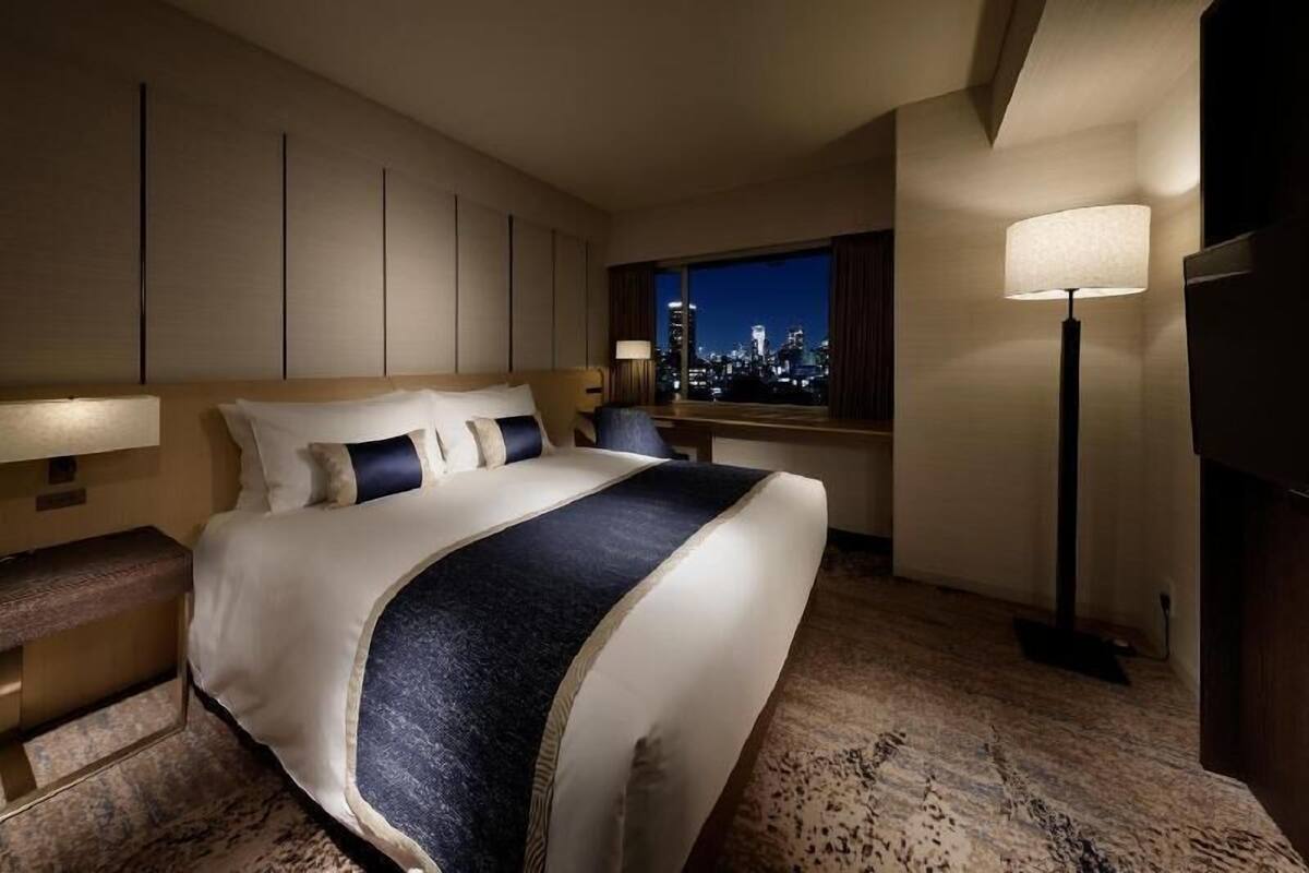 A hotel room with a large bed, a city view, a standing lamp, and a television.