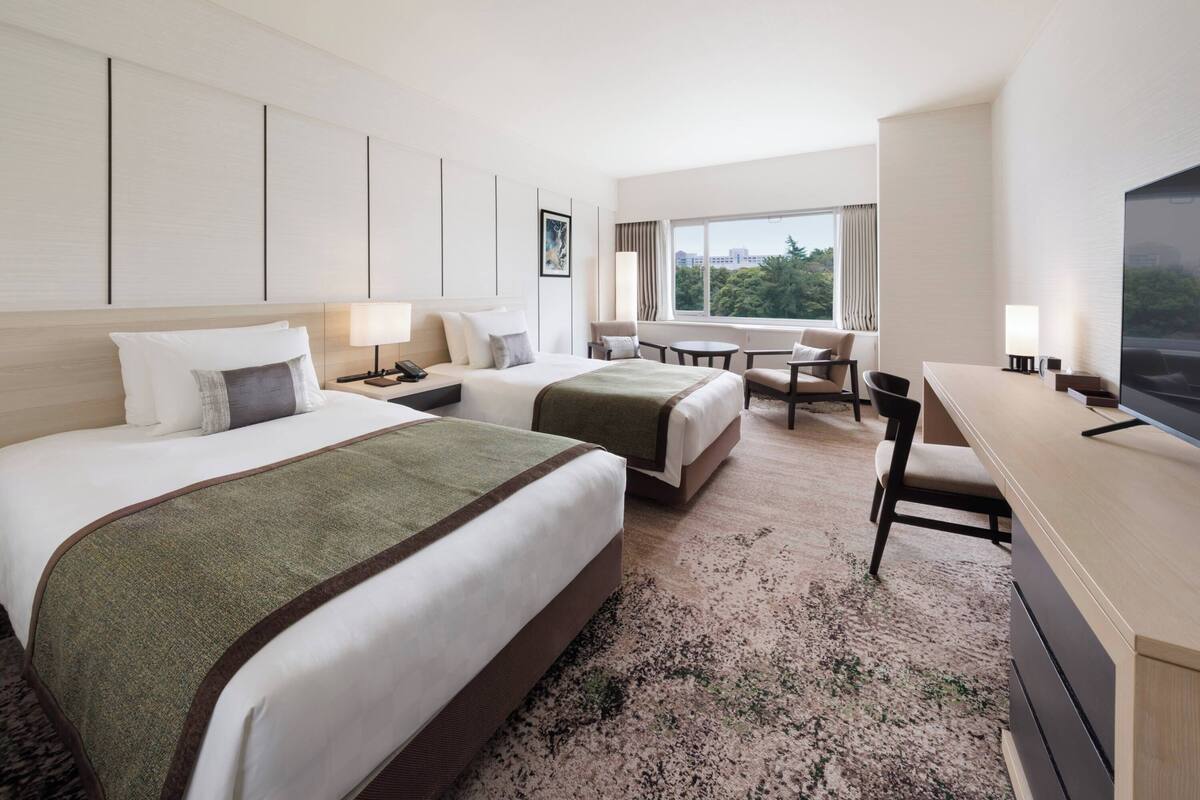A hotel room with two beds, a desk, a chair, a TV, and a window with a city view.