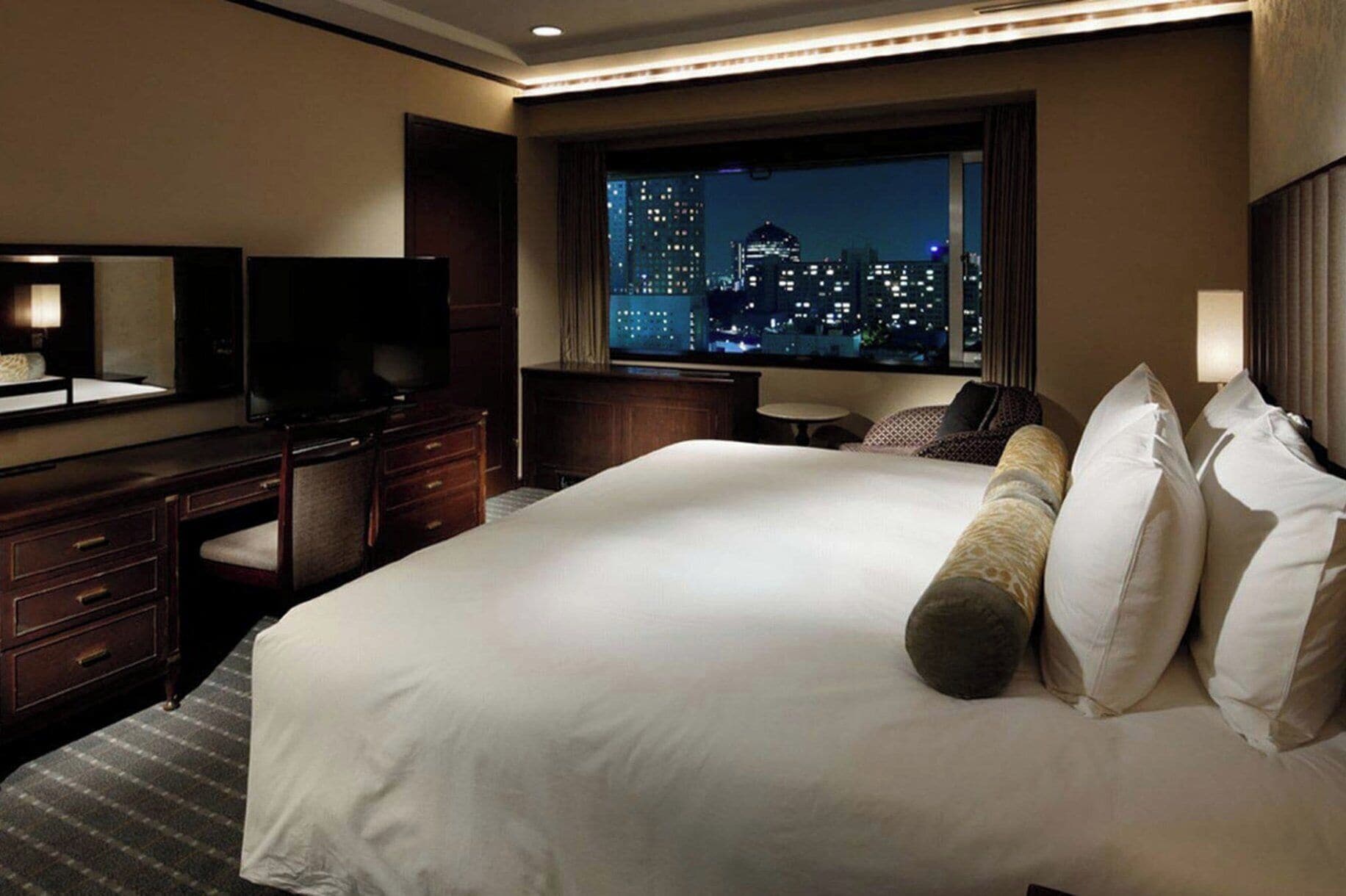 A hotel room with a large bed, a television, a desk, and a window with a city view at night.