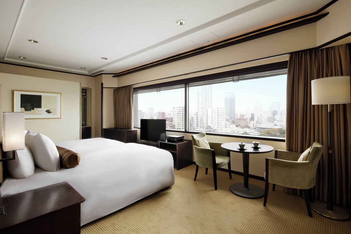 A hotel room with a large bed, a desk with a computer, a chair, a small table, and a large window with a city view.