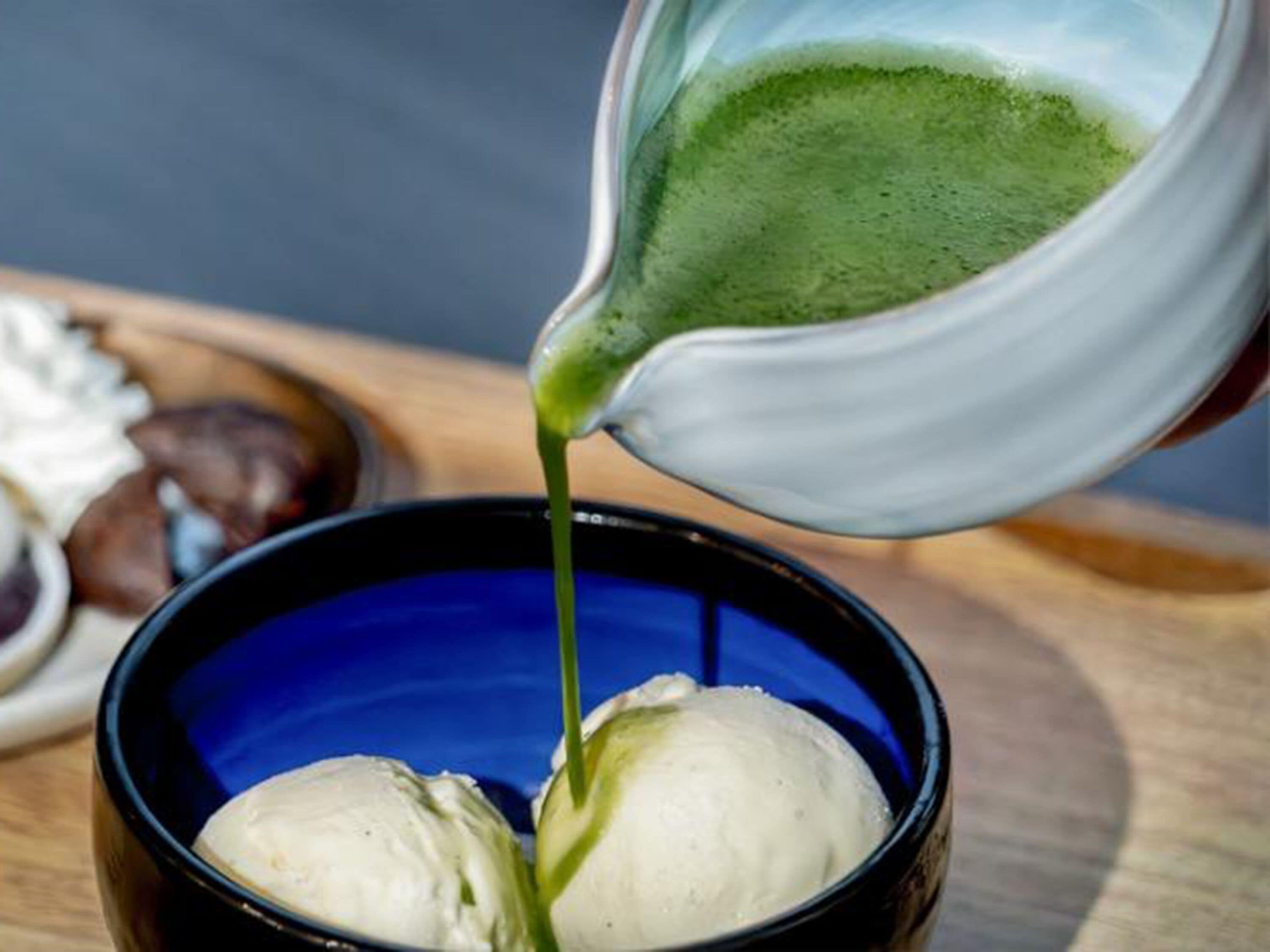 A cup of green tea being poured into a blue cup containing two scoops of ice cream.