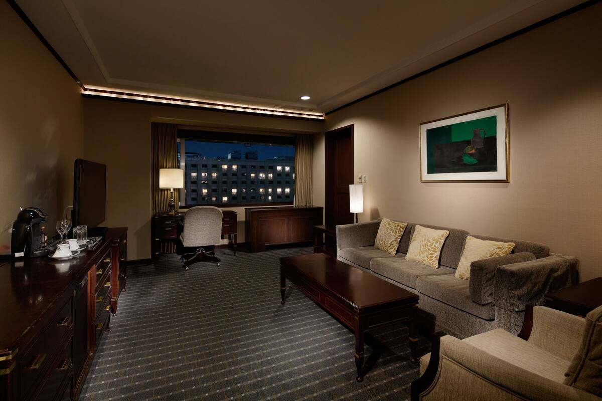 A hotel room with a sofa, armchairs, a desk, and a view of a cityscape at night.