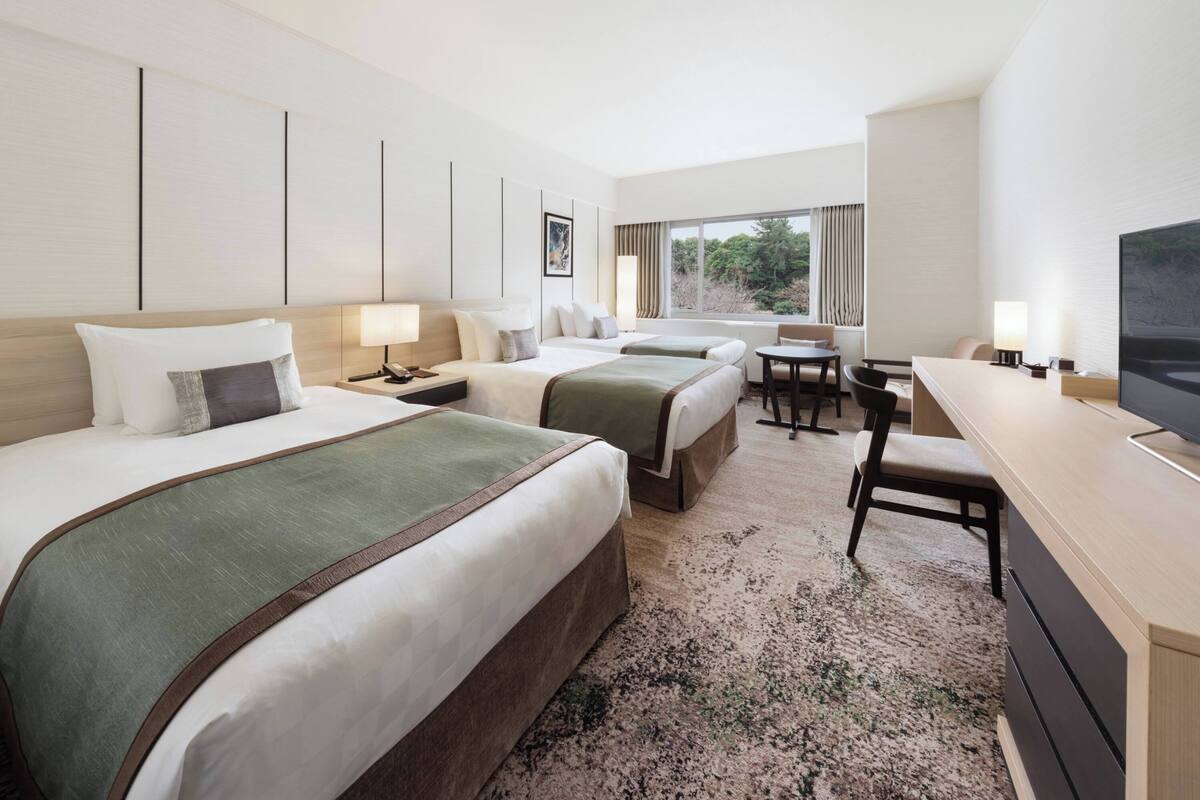 A hotel room with two beds, a desk, a chair, a TV, and a window with curtains.