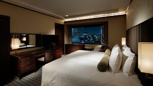 A hotel room with a large bed, a desk, a chair, a television, and a window with a city view.