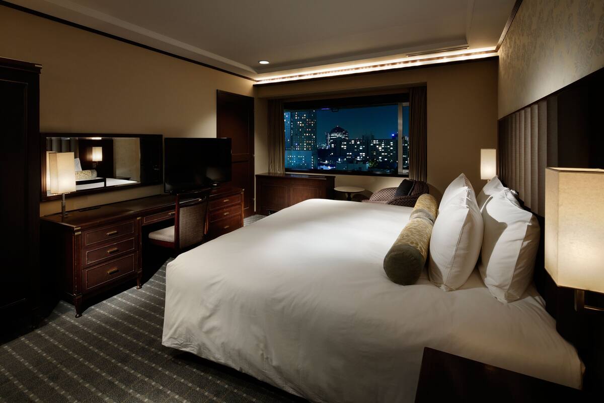 A hotel room with a large bed, a desk, a chair, a television, and a window with a city view.