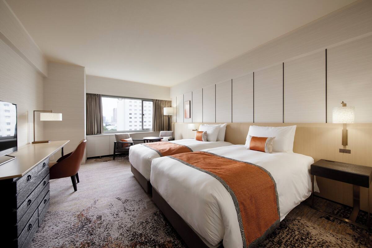 A hotel room with two beds, a desk, a chair, and a window with a city view.