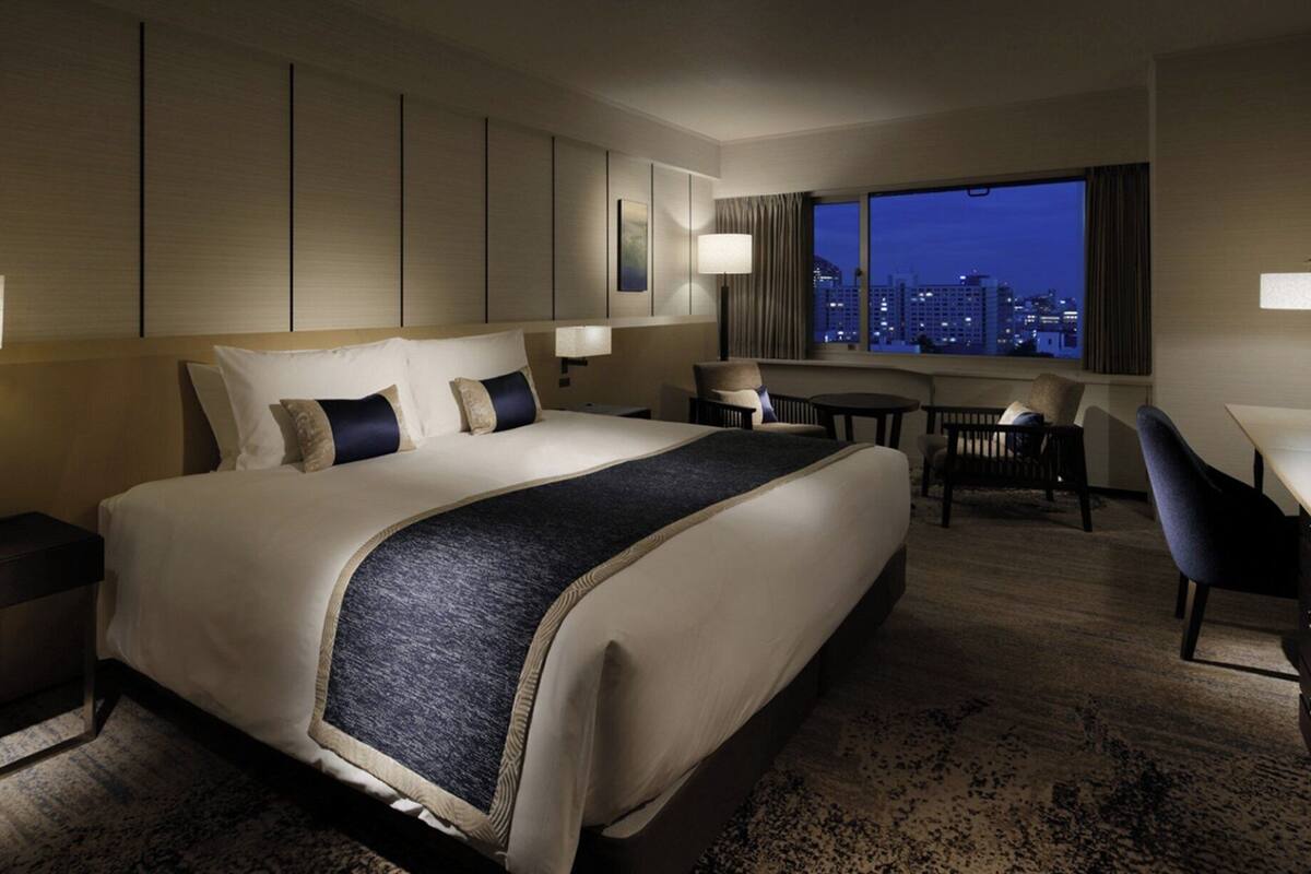 A hotel room with a large bed, a desk, a chair, and a view of the city at night.